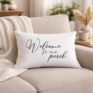 May include: A white rectangular pillow with the words "Welcome to our porch" in black script. The pillow is on a beige sofa, with a cream-colored throw blanket and decorative items in the background.