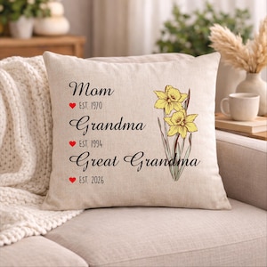 May include: A square beige cushion featuring the words "Mom," "Grandma," and "Great Grandma" in script. Each title has an establishment year and a red heart. A floral illustration of yellow daffodils is on the right side.