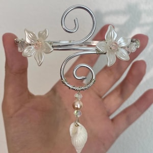 May include: A silver arm cuff with clear flower details and a seashell pendant. The cuff has a spiral design and is decorated with beads and crystals. The seashell is white.