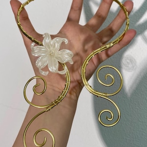 Peut inclure: Un collier en fil d'or avec un pendentif transparent en forme de fleur. Le collier présente un design circulaire avec des volutes décoratives et un accent floral. Le collier est tenu par une main.