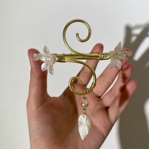 May include: A gold-toned metal bracelet with a floral design. The bracelet features two clear, flower-shaped accents and a dangling seashell pendant. The design includes a swirl and is held together with wire.
