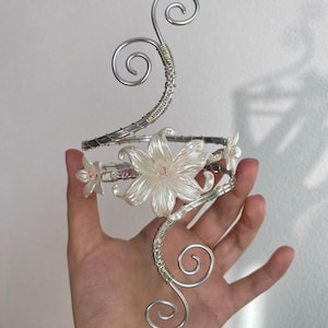Pode incluir: Uma pulseira de braço prateada com um design floral. A pulseira apresenta uma grande flor central e flores menores, com um design enrolado em arame e redemoinhos decorativos. As flores são brancas com um brilho perolado.