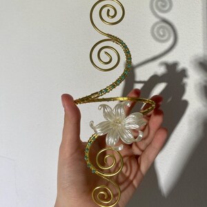 Peut inclure: Un bracelet de bras doré avec un motif floral. Le bracelet présente un design en spirale avec de petites perles turquoise et une fleur blanche. Le design est maintenu par un fin fil d'or.