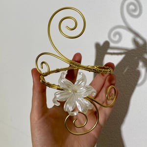 May include: A gold-toned metal arm cuff with a clear, flower-shaped centerpiece. The cuff features decorative swirls and is held in a hand. The flower has multiple petals and a clear, sparkling appearance. The metal has a shiny finish.