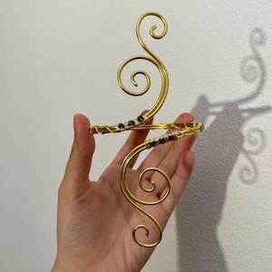 May include: A gold-colored arm cuff with a swirling design and small purple beads. The cuff features an open design with two curved sections connected by a band. The decorative piece is held in a hand against a white background.