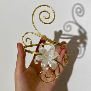 May include: Gold-toned arm cuff featuring a clear, flower-shaped centerpiece. The cuff has a spiral design and is made of wire. The flower has a crystal-like appearance. The cuff is designed to be worn on the upper arm.