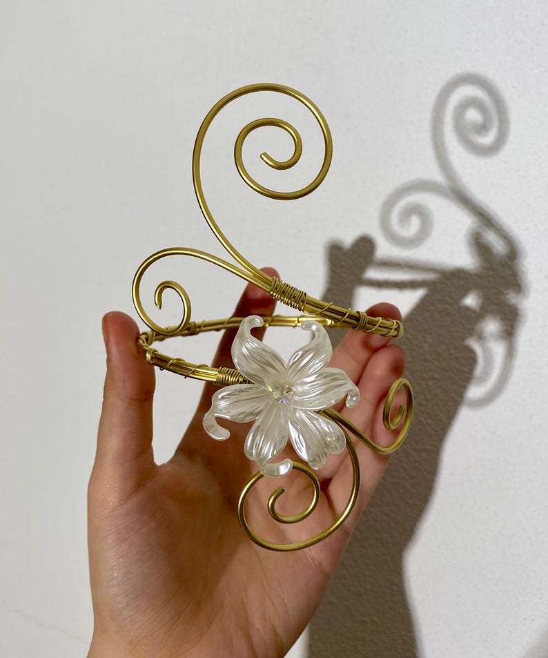 May include: Gold-toned arm cuff featuring a clear, flower-shaped centerpiece. The cuff has a spiral design and is made of wire. The flower has a crystal-like appearance. The cuff is designed to be worn on the upper arm.