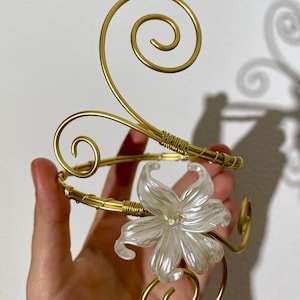 May include: A gold-toned arm cuff with a floral design. The cuff features a large, pearlescent white flower and decorative swirls. The metal is wrapped in gold wire in several places.