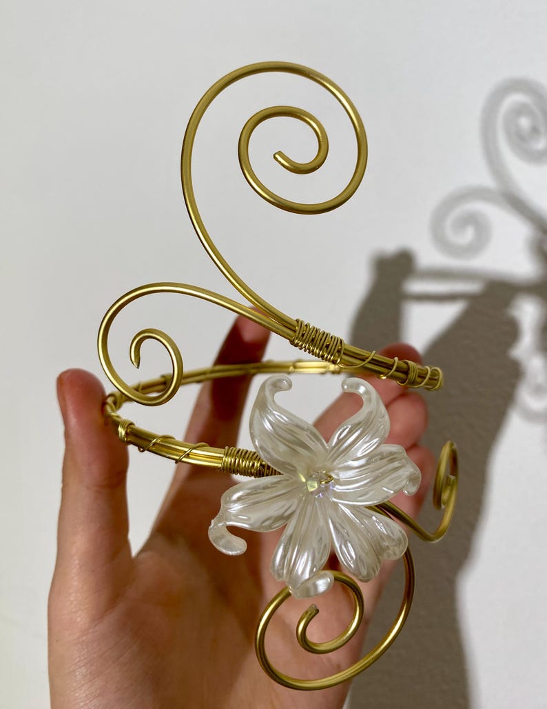May include: A gold-toned arm cuff with a floral design. The cuff features a large, pearlescent white flower and decorative swirls. The metal is wrapped in gold wire in several places.
