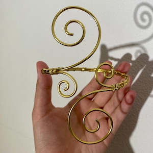 Può includere: Un bracciale dorato con un design a spirale aperto. Il bracciale presenta molteplici accenti a spirale ed è tenuto in mano su uno sfondo bianco. Il metallo sembra essere simile a filo, con una finitura lucida.