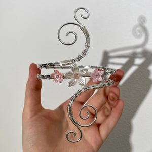 Può includere: Un bracciale argentato con un design floreale. Il bracciale presenta tre fiori rosa e trasparenti e riccioli decorativi. Il metallo è avvolto attorno al braccio in un design a spirale. Il bracciale è progettato per essere indossato sulla parte superiore del braccio.