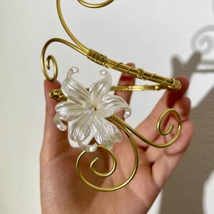 May include: A gold-colored metal decorative arm cuff with a clear, white flower accent. The cuff features a swirling design and is held in a hand. The flower has multiple petals and is centered on the cuff.