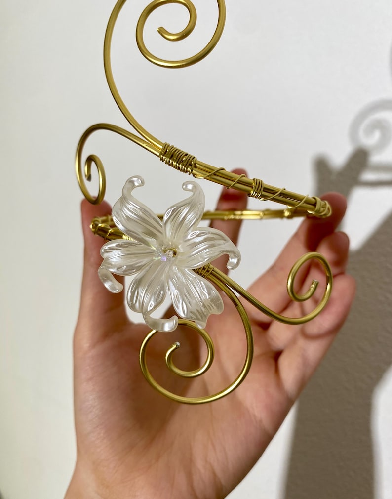 May include: A gold-colored metal decorative arm cuff with a clear, white flower accent. The cuff features a swirling design and is held in a hand. The flower has multiple petals and is centered on the cuff.