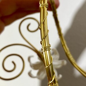 May include: Close-up of a gold-colored decorative object with a floral design. The object has a curved shape with intricate wire detailing and a white flower accent. The design is held by a hand.