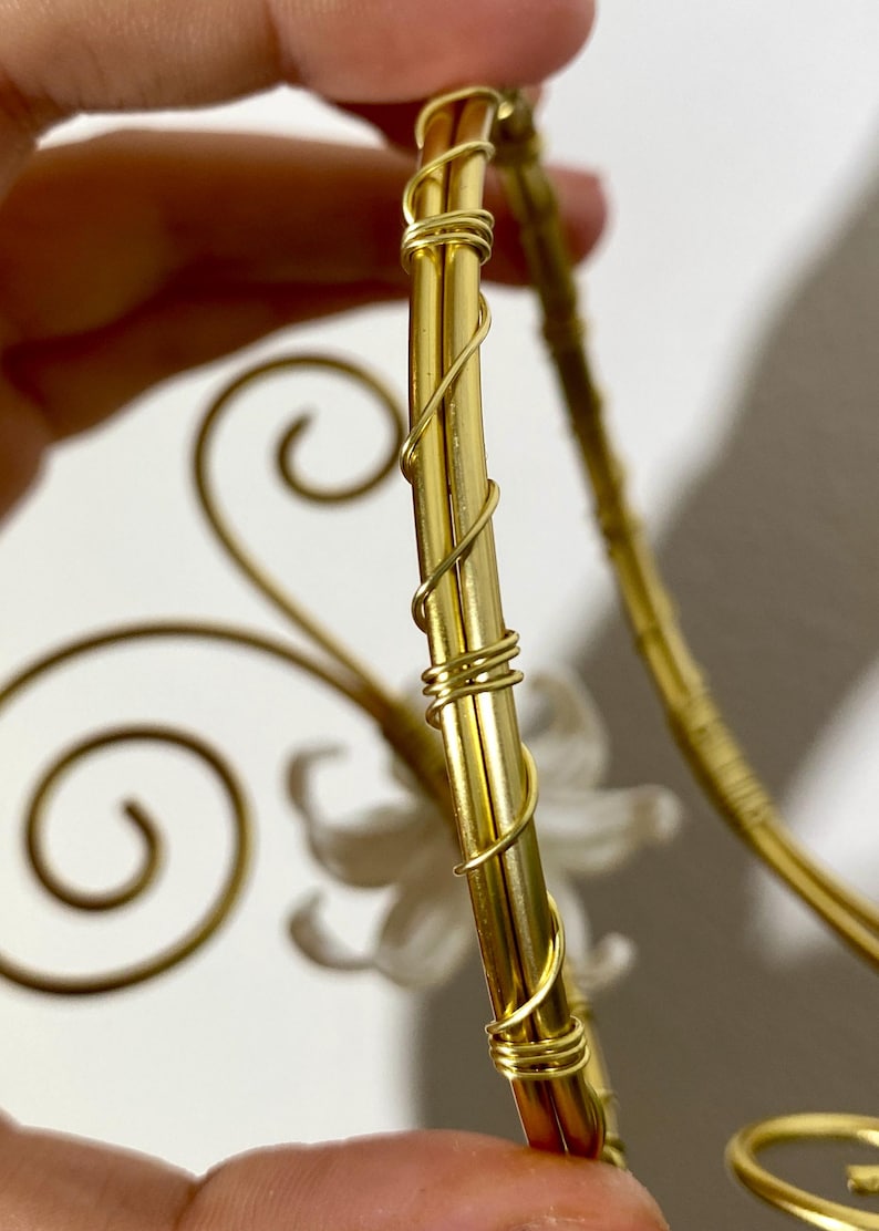 May include: Close-up of a gold-colored decorative object with a floral design. The object has a curved shape with intricate wire detailing and a white flower accent. The design is held by a hand.