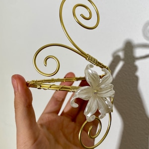 May include: A gold-colored arm cuff with a floral accent. The cuff features a delicate design with a white flower and swirling accents. The metal is wrapped in a wire pattern. The overall design is elegant and decorative.