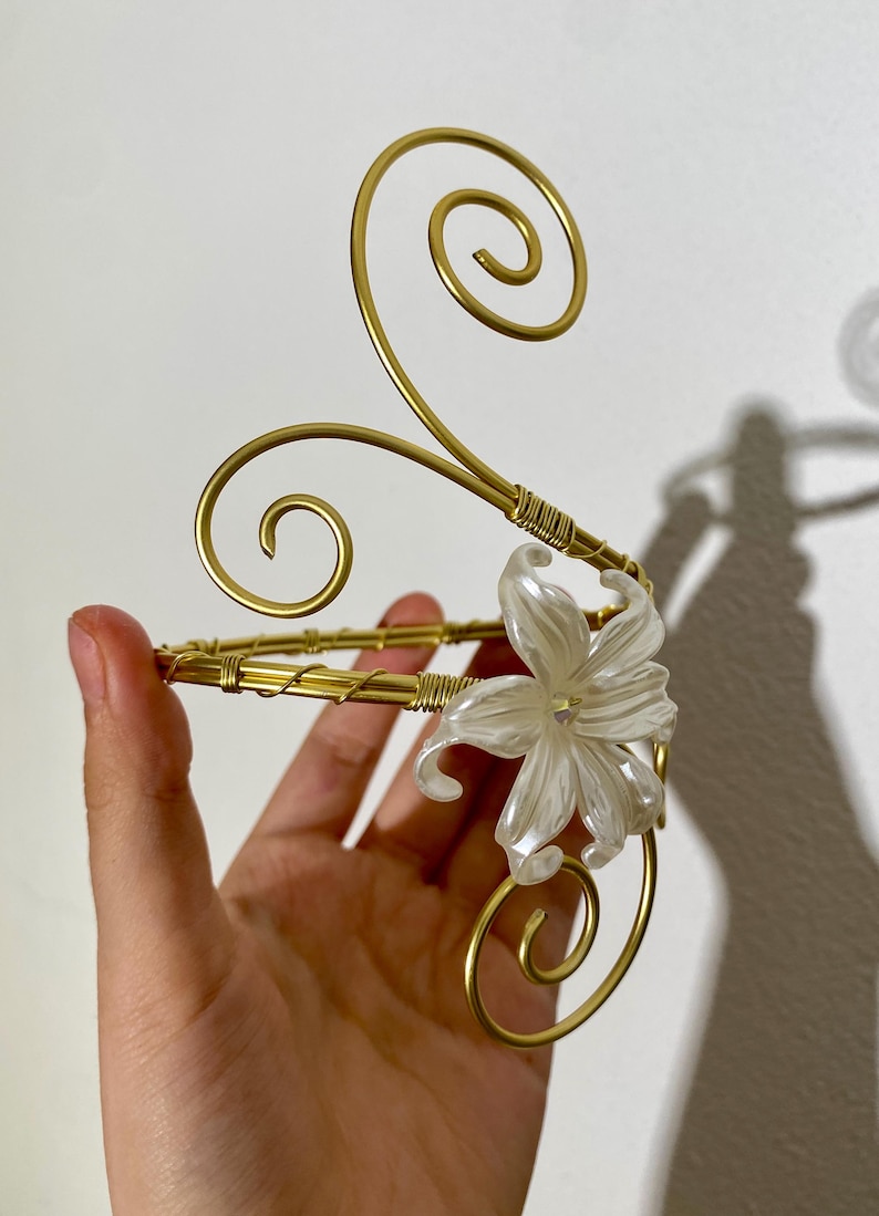 May include: A gold-colored arm cuff with a floral accent. The cuff features a delicate design with a white flower and swirling accents. The metal is wrapped in a wire pattern. The overall design is elegant and decorative.
