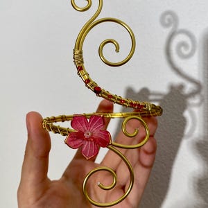 Può includere: Un bracciale dorato con un design floreale. Il bracciale presenta un fiore rosso ed è adornato con perline rosse. Il design include elementi decorativi a spirale, creando un accessorio elegante e artistico.