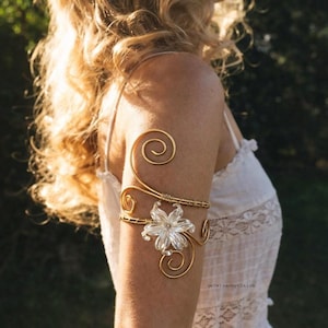 May include: A gold arm cuff with a floral design and spiral detailing. The cuff has a clear, flower-shaped centrepiece and is worn on the upper arm. The jewellery is set against a blurred background.