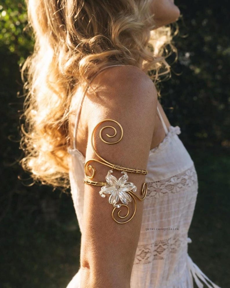 May include: A gold-colored arm cuff with a floral design and spiral accents. The cuff features a clear, flower-shaped centerpiece and is worn on the upper arm. The jewelry is set against a blurred background.