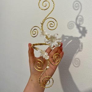 May include: A gold-colored wire arm cuff with a floral design. The cuff features delicate white flowers and pearl accents. The wire is shaped into elegant swirls and curves, creating a decorative accessory.