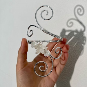 Op de afbeelding: Een zilverkleurig metalen decoratief haaraccessoire met een bloemenontwerp. Het accessoire heeft een gebogen, open ontwerp met wervelende accenten en kleine witte bloemen. Het stuk wordt in een hand gehouden tegen een effen witte achtergrond.