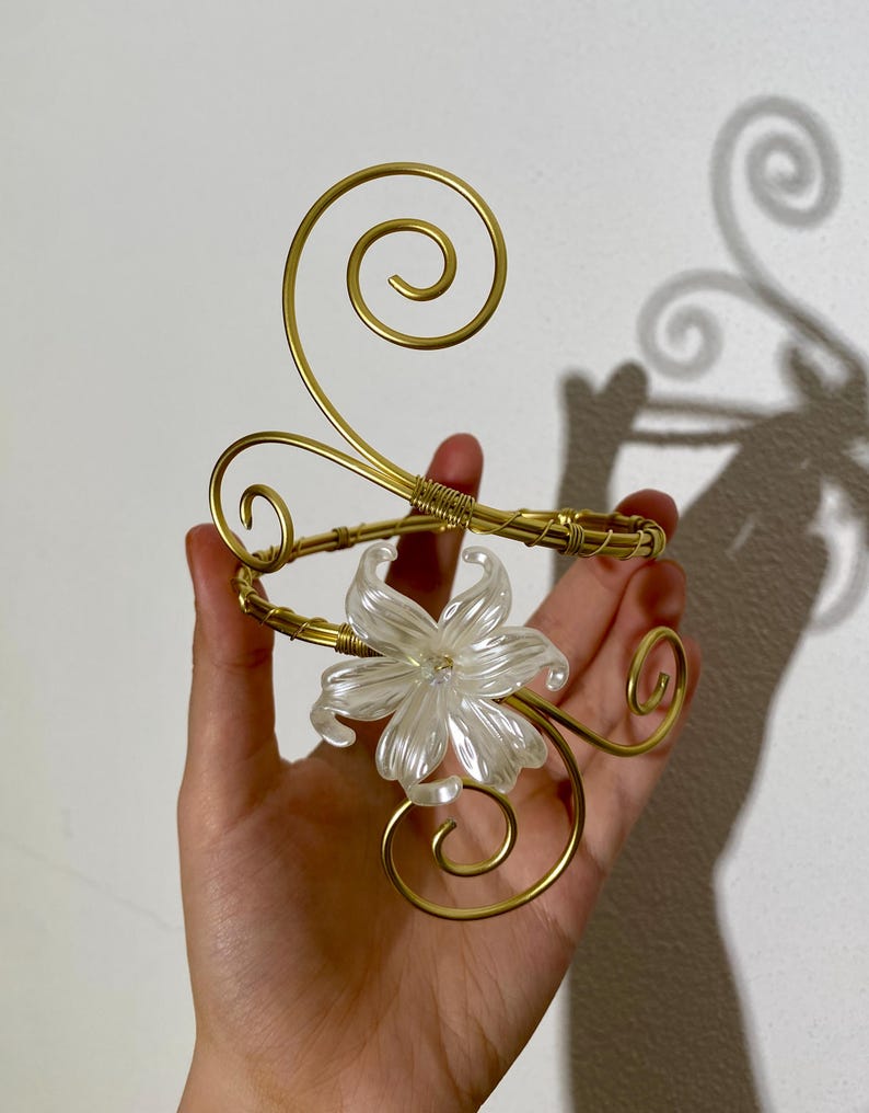 May include: A gold-toned metal arm cuff with a clear, flower-shaped centerpiece. The cuff features decorative swirls and is held in a hand. The flower has multiple petals and a clear, sparkling appearance. The metal has a shiny finish.