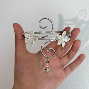 Peut inclure: Un bracelet de bras argenté avec un motif floral. Le bracelet comporte deux accents de fleurs blanches et de petites perles bleues. Un petit charme de fleur pend en bas. Le bracelet est tenu dans une main.