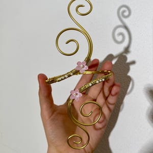 May include: A gold-coloured arm cuff with a floral design. The cuff features a swirling wire design with pink flower accents and small white beads. The cuff is held up against a white background.