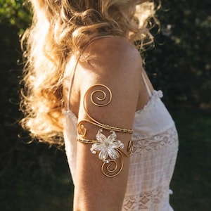 Pode incluir: Um bracelete de arame em tom dourado com um detalhe floral central transparente de várias pétalas. O arame metálico é moldado em espirais decorativas que se estendem acima e abaixo do elemento floral central, envolvendo a parte superior do braço.