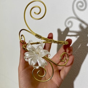 May include: A gold-toned arm cuff with a floral accent. The cuff features a delicate, swirling wire design and a clear, flower-shaped embellishment. The cuff is held in a hand against a white background.