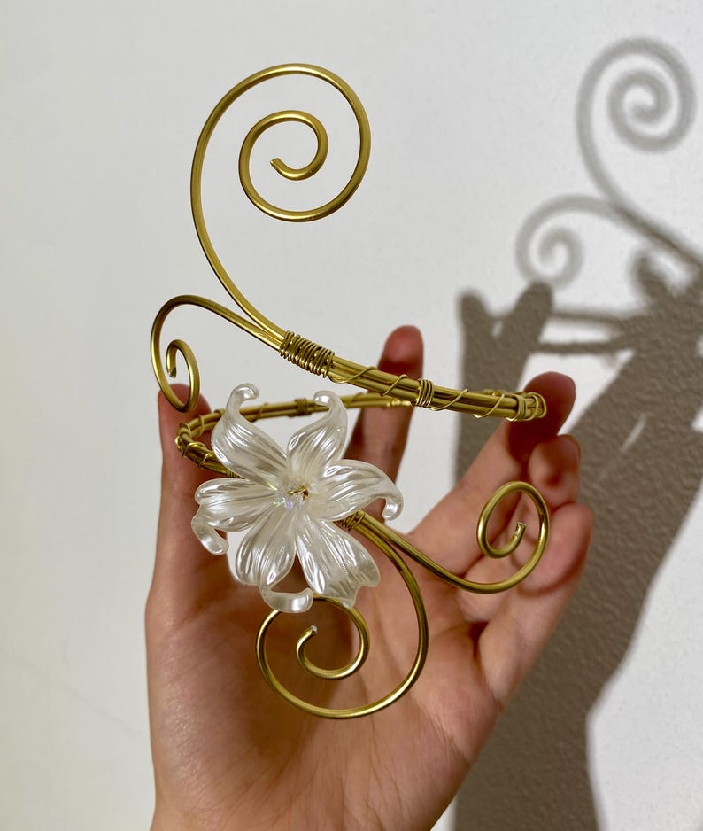 May include: A gold-toned arm cuff with a floral accent. The cuff features a delicate, swirling wire design and a clear, flower-shaped embellishment. The cuff is held in a hand against a white background.