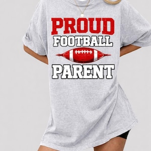 Puede incluir: Una camiseta gris claro con las palabras "PROUD FOOTBALL PARENT" en letras rojas y blancas. Un gráfico de fútbol americano rojo y blanco está centrado entre las palabras. La camiseta es de corte holgado.