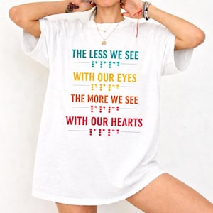 May include: White t-shirt with the text "THE LESS WE SEE WITH OUR EYES THE MORE WE SEE WITH OUR HEARTS" in teal, yellow, orange, and red. Braille dots are below each line of text. The shirt is a casual, everyday wear item.