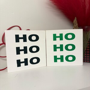 Puede incluir: Dos bloques de madera blancos con el texto "HO HO HO" en negro y verde. Los bloques se muestran sobre una superficie blanca, con elementos decorativos rojos en el fondo. Una decoración festiva navideña.