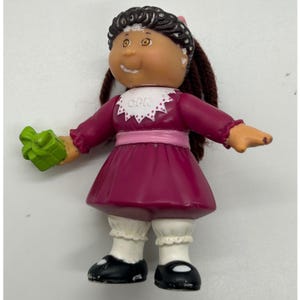 May include: A small plastic doll with dark brown yarn hair, wearing a magenta dress with a white collar featuring the letters GPK, a light pink waistband, white bloomers, and black shoes. The doll holds a small, bright green gift box in one hand.