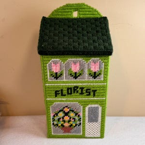 May include: A green, handmade needlepoint wall hanging shaped like a building, with a dark green roof and the word "FLORIST" across the front. The design includes pink tulips and a flower basket.