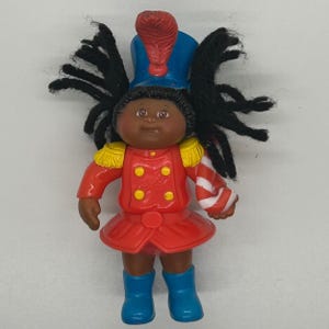 May include: A small plastic doll with dark skin and black yarn hair, wearing a blue hat with a red feather, a red jacket with yellow epaulets and buttons, a red skirt, and blue boots. The doll holds a red and white striped candy cane accessory.