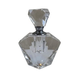 Puede incluir: Un frasco de perfume de vidrio transparente facetado, con un cuerpo en forma de diamante y un tapón geométrico. El frasco tiene un collar de metal plateado. El diseño es elegante y vintage, adecuado para exhibición o uso.