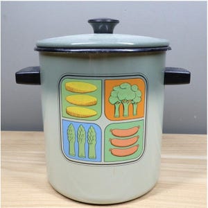 Może przedstawiać: W vintage jasnozielonym emaliowanym garnku z pokrywką i czarnymi uchwytami. Garnek posiada kolorową grafikę kukurydzy, brokułów, szparagów i kiełbasek. Garnek jest prawdopodobnie używany do gotowania.