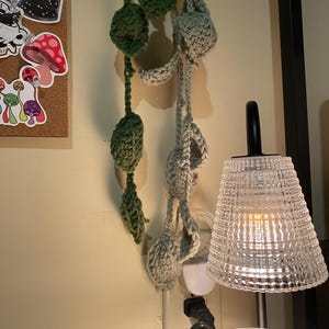 Może przedstawiać: Dekoracja ścienna wykonana z zielonych i szarych, dzianych elementów w kształcie roślin, wisząca z sufitu. Dekoracja wisi w pobliżu białego gniazdka elektrycznego i lampy z przezroczystym kloszem.