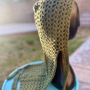 May include: A green crochet hooded scarf with a loose, open weave. The scarf is draped over a mannequin head and has long ties.