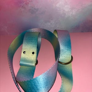 May include: A multi-coloured belt in shades of blue, yellow, and purple. It features a metal buckle and a metal ring. The belt is made from a smooth, woven fabric.