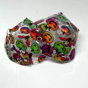 Könnte beinhalten: Ein klarer, gebogener Gegenstand mit einem Halloween-Muster. Das Design zeigt bunte Kürbisse, Fledermäuse und Sterne. Das Accessoire ist mit silbernen Ösen befestigt und ist wahrscheinlich ein dekorativer Artikel.