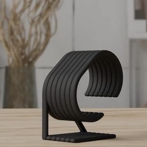 May include: Black, wave-shaped, decorative stand made of multiple stacked, thin, horizontal pieces. The stand is designed to hold a watch or other small object.