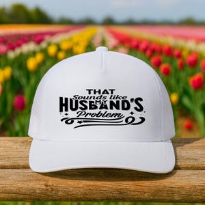 May include: White trucker hat with a mesh back and a curved brim. The front of the hat features the text "THAT Sounds like MY HUSBAND'S Problem" in black script and block letters. The hat is set against a blurred background of colorful flowers.
