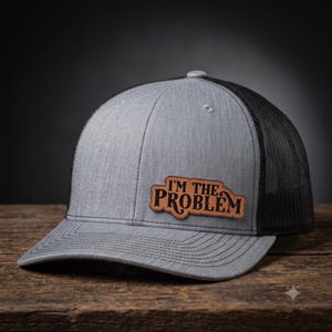 May include: A grey heather baseball cap with a black mesh back and a brown leather patch on the front. The patch features the text I'M THE PROBLEM in a dark, stylized font. The cap has a structured crown and a curved brim with stitching details.