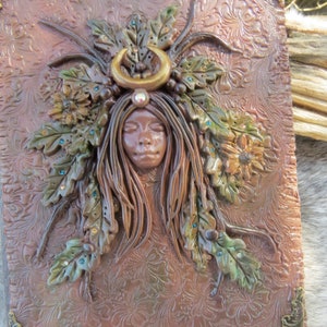 May include: A brown leather journal with a sculpted design of a woman's face surrounded by leaves and flowers. The woman has her eyes closed and is wearing a crescent moon headband.
