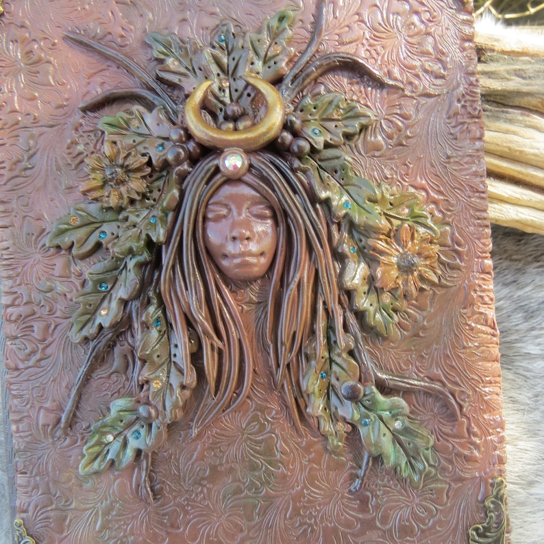 May include: A brown leather journal with a sculpted design of a woman's face surrounded by leaves and flowers. The woman has her eyes closed and is wearing a crescent moon headband.