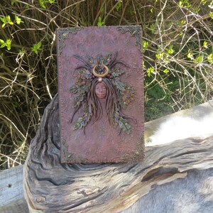 May include: A brown leather journal with a decorative embossed design. The cover features a sculpted image of a woman with a crown of leaves and a crescent moon.